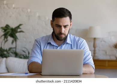 Focused millennial business man using laptop at table. Young entrepreneur, freelancer in casual, employee, worker using compute at home office workplace, typing, chatting online Foto Stock