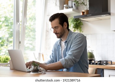 Focused millennial Caucasian man sit at table at home kitchen look at laptop screen working distant. Young male use computer, texting or messaging online on device. Communication, technology concept. - Powered by Shutterstock