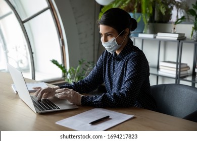 Focused Millennial Female Indian Company Employee Worker Manager In Protective Facial Mask And Medical Gloves Working On Laptop At Workplace, Keeping Covid 19 Dangerous Virus Quarantine Measures.