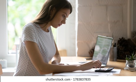Focused millennial female sit at desk pay bills taxes online on laptop, calculate finances on machine, concentrated young woman manage household family budget expenditures on calculator