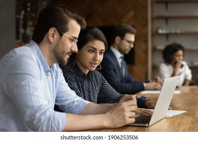 Focused millennial male intern look at laptop screen listen to skilled indian female mentor. Hindu woman experienced worker consult young man new employee help in computer work at corporate network