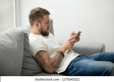 Focused Millennial Man Sitting On Cozy Couch Holding Tablet Reading Online News Or Checking Mail, Lazy Guy Relaxing On Sofa Playing App Games Or Downloading New Applications, Male Browsing Web