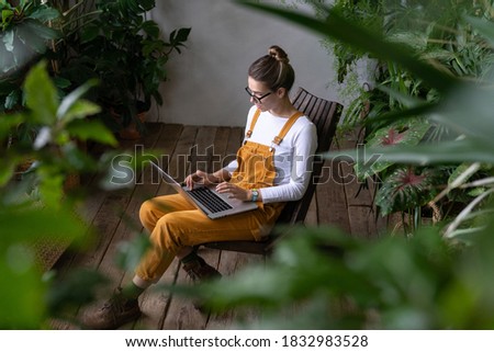 Focused millennial woman gardener in overalls watching educational webinar on laptop, writing blog or email, studying, remotely online work in her home garden surrounded by tropical plants. E-learning
