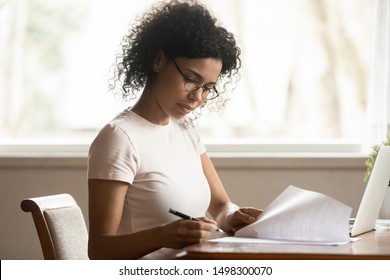 Focused mixed race ethnicity millennial businesswoman in glasses work at laptop read sign paper documents, concentrated african American woman student consider paperwork do corrections