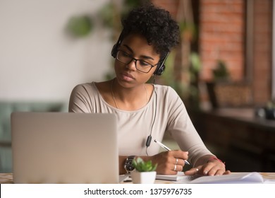 Focused mixed race woman wearing headphones watching webinar write notes study online with online teacher, african female student learning language computer course on laptop listen translate lecture