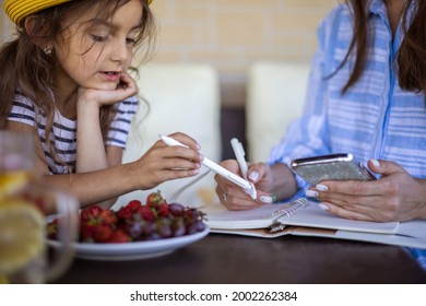 Focused Mother And Daughter Looking Paper Schedule Notebook Together Relaxing At Outdoor Summer Terrace. Confident Family Planning Watching Data At Table With Seasonal Flowers And Fruits, Drinks