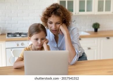 Focused Mother With Little Daughter Looking At Laptop Screen Together Studying Or Watching Webinar Sitting At Table In Kitchen, Pensive Mom And Preschool Girl Kid Pondering Problem With Device