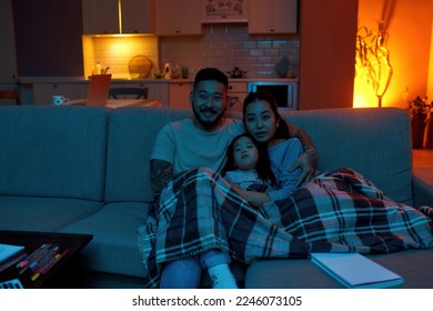 Focused mother, smiling father and little daughter watching TV or movie under blanket on sofa at home. Asian family relationship. Parenthood. Domestic entertainment and leisure. Evening or night - Powered by Shutterstock