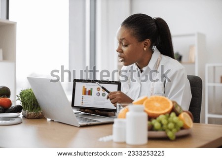 Focused multicultural expert with meal plan on tablet conducting web meeting using portable laptop at work. Skilful female nutritionist explaining details of slimming program for client online.