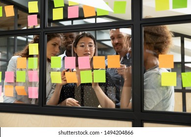 Focused multiethnic business team people brainstorm on corporate project together write strategy tasks on post in sticky notes stand behind glass wall, diverse staff group planning management concept