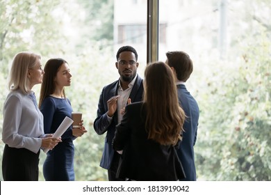 Focused Multiracial Businesspeople Gather In Office Near Window Talk Discuss Business Ideas In Group Together. Multiethnic Diverse Colleagues Coworkers Brainstorm At Casual Meeting. Teamwork Concept.