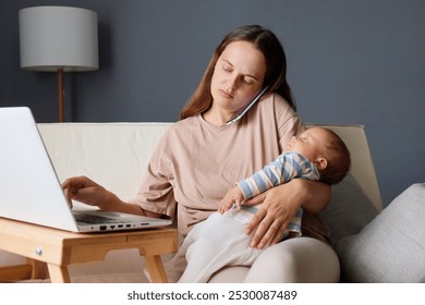 Focused multitasking woman handling phone call working on her laptop and caring for her baby typing on keyboard balancing freelance work and motherhood while sitting on couch - Powered by Shutterstock