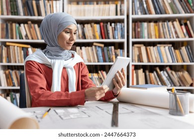 A focused Muslim woman in a hijab using a digital tablet at a library table with architectural blueprints and books around. - Powered by Shutterstock