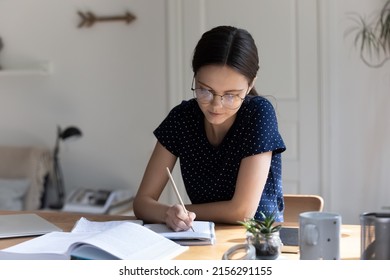 Focused nerdy student girl wearing glasses, writing essay in copy book at workplace table, making notes, summary from open book, doing school, college home task, research study - Powered by Shutterstock