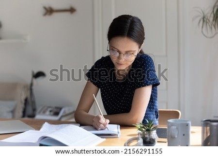 Focused nerdy student girl wearing glasses, writing essay in copy book at workplace table, making notes, summary from open book, doing school, college home task, research study