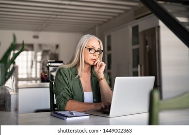 Focused Old Woman At Home Using Laptop. Senior Stylish Entrepreneur With Notebook And Pen Wearing Eyeglasses Working On Computer. Serious Lady Analyzing And Managing Domestic Bills And Home Finance.