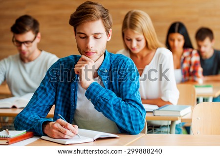 Focused on exam. Group of concentrated young students writing something in their note pads while sitting at their desks in the classroom