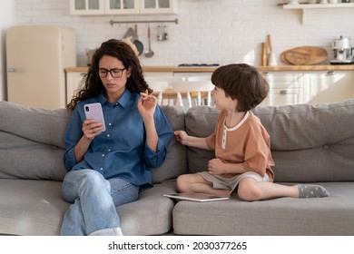 Focused on mobile phone woman working mother pay no attention on bored small kid. Young mom or nanny texting in smartphone sitting on sofa next to little son need parent love care and communication