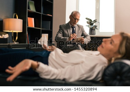 Focused on writing. Serious concentrated grey-haired psychotherapist writing while sitting in a chair in front of his female patient