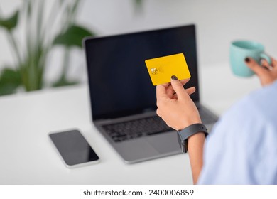 Focused on yellow credit card in hand on laptop screen, online shopping or banking, with phone and drink cup of coffee. Digital banking and online shopping, technology and money