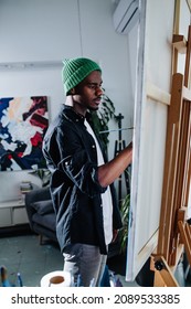 Focused Peaceful Black Man Painting On An Easel Inside Of His Messy Disorderly Apartment. An Abstract Painting On The Wall Behind.