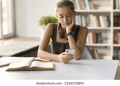 Focused pensive hipster student girl typing on mobile phone, using learning application, educational service in college s library, texting message on cellphone, sitting at open books - Powered by Shutterstock