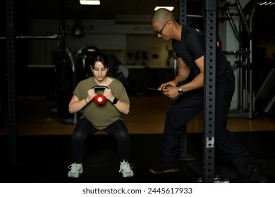 Focused personal trainer with tablet in hand supervises a female client as she squats holding a kettlebell - Powered by Shutterstock