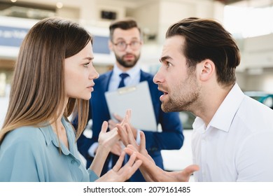 Focused Photo Of Confused Male Shop Assistant Looking At Angry Arguing Young Family Caucasian Couple While They Are Deciding Which Car Auto To Choose Buy. Expensive High Price.