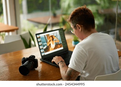 Focused photo editor with tattoos retouches images on laptop at coworking space. Professional with camera reviews creative visual content. Freelancer works in modern office, digital workflow. - Powered by Shutterstock