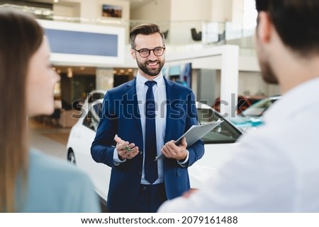 Focused photo of young male shop assistant explaining talking offering buying new car to young caucasian family husband and wife at auto dealer shop.