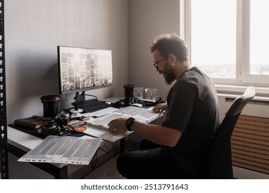 A focused photographer analyzes film negatives in a well-lit room, surrounded by photography equipment and a large monitor. - Powered by Shutterstock