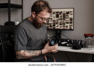 Focused photographer with tattoos inspects his camera lens in a well-organized workspace with photography equipment. - Powered by Shutterstock