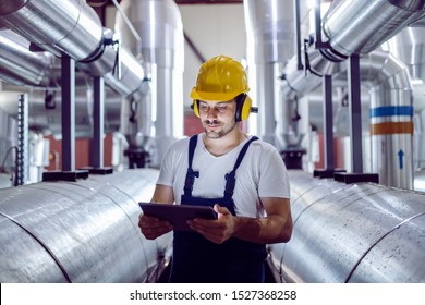 Focused Plant Worker In Overalls, With Protective Helmet On Head And Antiphons On Ears Using Tablet For Checking Machine.