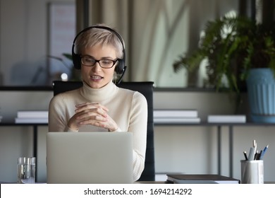 Focused pleasant 30s businesswoman in eyeglasses wearing wireless headphones with microphone, looking at laptop screen, holding video call negotiations meeting online with partners alone in office. - Powered by Shutterstock