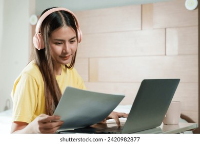 Focused and poised, young woman with headphones examines document, multitasking in work from home. female reviews paperwork with attention,  suggesting a backdrop of music during work with labtop - Powered by Shutterstock