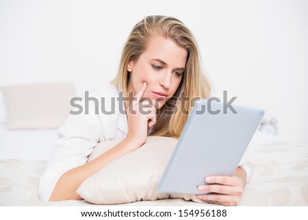 Focused pretty model using her tablet lying on cosy bed in bright bedroom