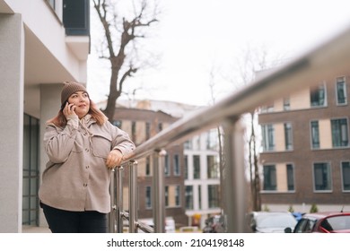 Focused Pretty Overweight Woman In Warm Hat And Jacket Talking On Mobile Phone Standing Near Railing At City Street In Cloudy Autumn Day. Serious Obese Female Having Call On Smartphone Outdoors.