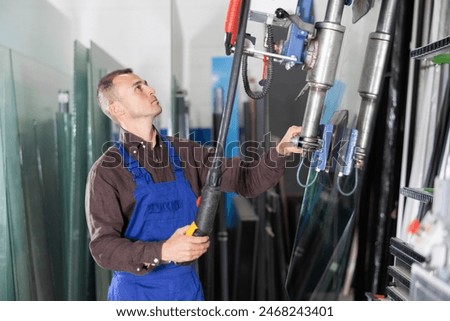 Focused professional operator maneuvering large glass sheets with vacuum lifter in industrial processing workshop