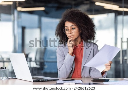 Focused project manager with curly hair holding document and looking with hesitation at computer screen. Suspicious woman checking accuracy of printed report and comparing with electronic version.