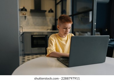 Focused redhead 10 years old boy with freckles looking to laptop screen and typing doing homework sitting at table on background of kitchen. Cute teen boy surfing internet on notebook computer at home - Powered by Shutterstock