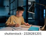 Focused redhead pupil student boy studying at home writing in exercise book doing homework, learning sitting at table under light of lamp at night. Clever schoolkid reading textbook sitting at desk.