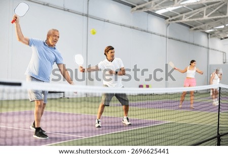 Focused resolved elderly man playing friendly pickleball match in team with male partner on indoors court. Concept of concentration in competition