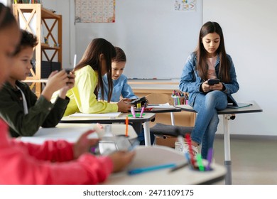 Focused schoolchildren using cellphones while sitting at desks in school classroom interior during break, copy space - Powered by Shutterstock