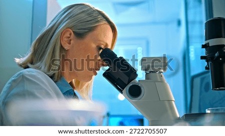 A focused scientist examines a sample under a microscope in a modern, high-tech lab. Blue lighting enhances the professional atmosphere, highlighting the precision required in medical research.