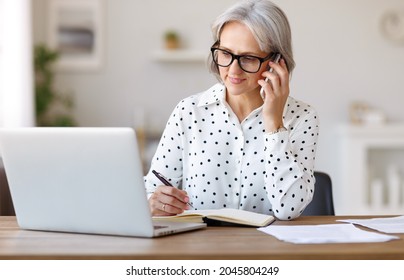 Focused senior business woman wearing glasses talking on mobile phone using laptop while working remotely at home, elderly woman holding smartphone and speaking with client at her workplace - Powered by Shutterstock