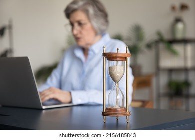 Focused senior gray haired business woman using laptop at hourglass on table, working on project, keeping schedule, completing tasks before deadline. Time management concept - Shutterstock ID 2156185843