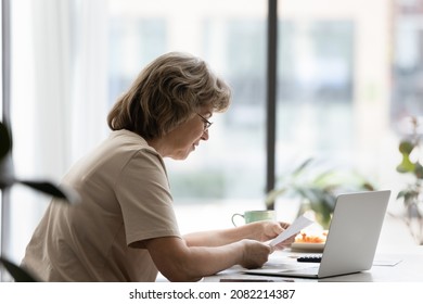 Focused Senior Homeowner, Landlady Reading Paper Document At Laptop, Doing Domestic Paperwork At Home, Paying Paper Bills, Invoices, Insurance Fees Online On Banking App, Sitting At Table. Side View