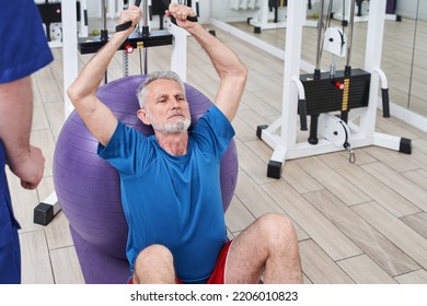 Focused Senior Man Laying On A Fitness Ball In Physical Rehabilitation Therapy With His Trainer