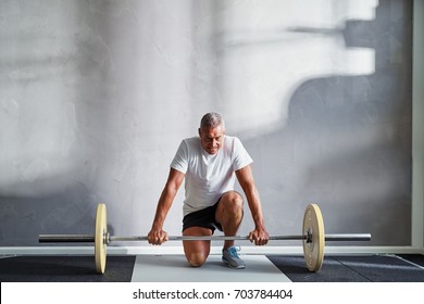 Focused Senior Man In Sportswear Kneeling Alone In A Gym Preparing To Lifting Weights During A Workout