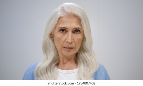 Focused senior woman looking camera inside. Serious old lady wagging finger on grey studio background. Portrait of disappointed female person gesticulating indoors.